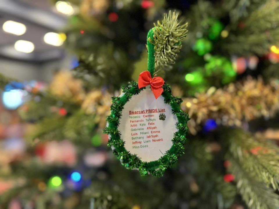 A festive ornament hanging on a Christmas tree with text that says "Bearcat PRIDE List" and student names listed below. The ornament has green garland trim and a red bow.