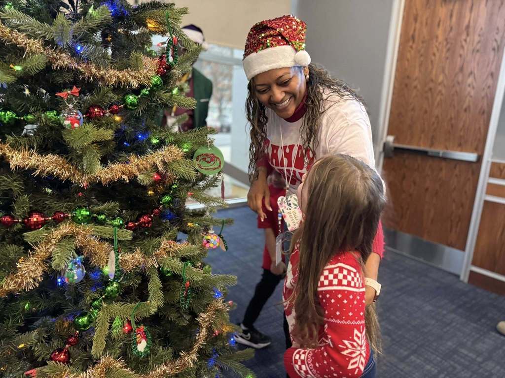 A district staff member wearing a Santa hat is interacting with a young girl near a decorated Christmas tree adorned with colorful ornaments and tinsel.
