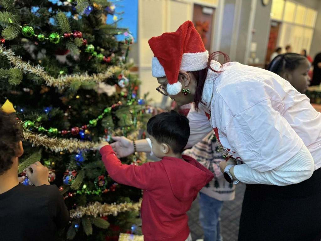 A district staff member wearing a Santa hat is helping a young child decorate a Christmas tree with tinsel and ornaments. Other children can be seen nearby and in the background.
