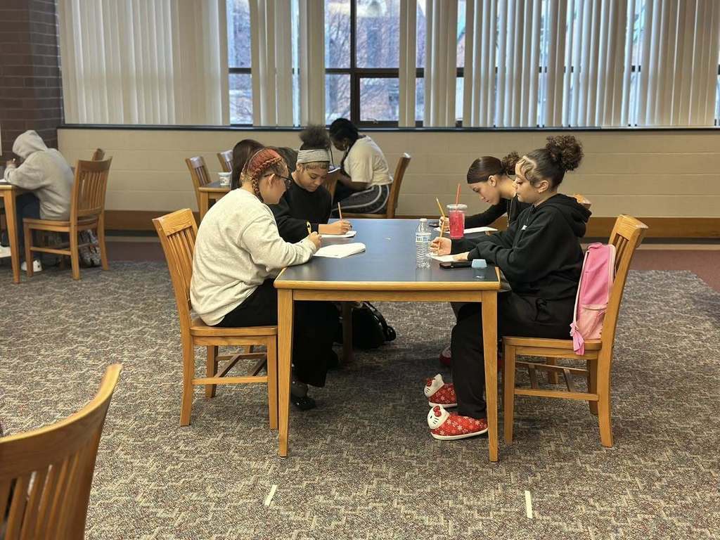 Students are sitting around a table in a school library, focused on writing and studying. Books and drinks can be seen on the table.