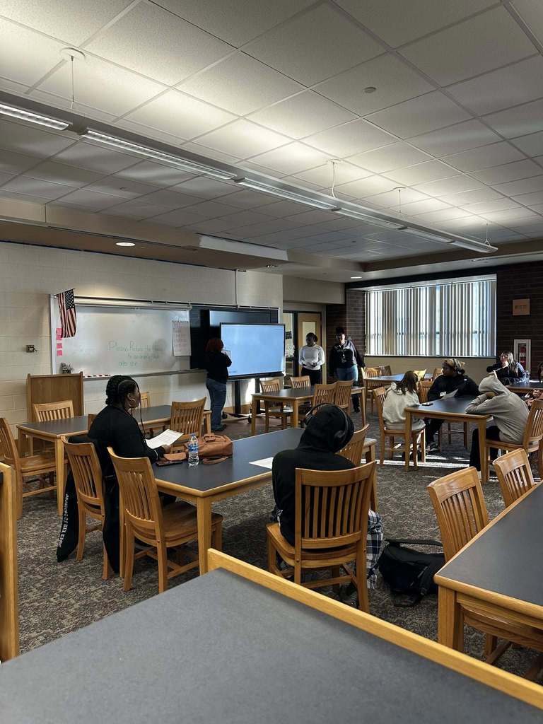 A school library filled with students who are sitting at wooden tables, facing a screen where two student presenters are standing near a smart board.