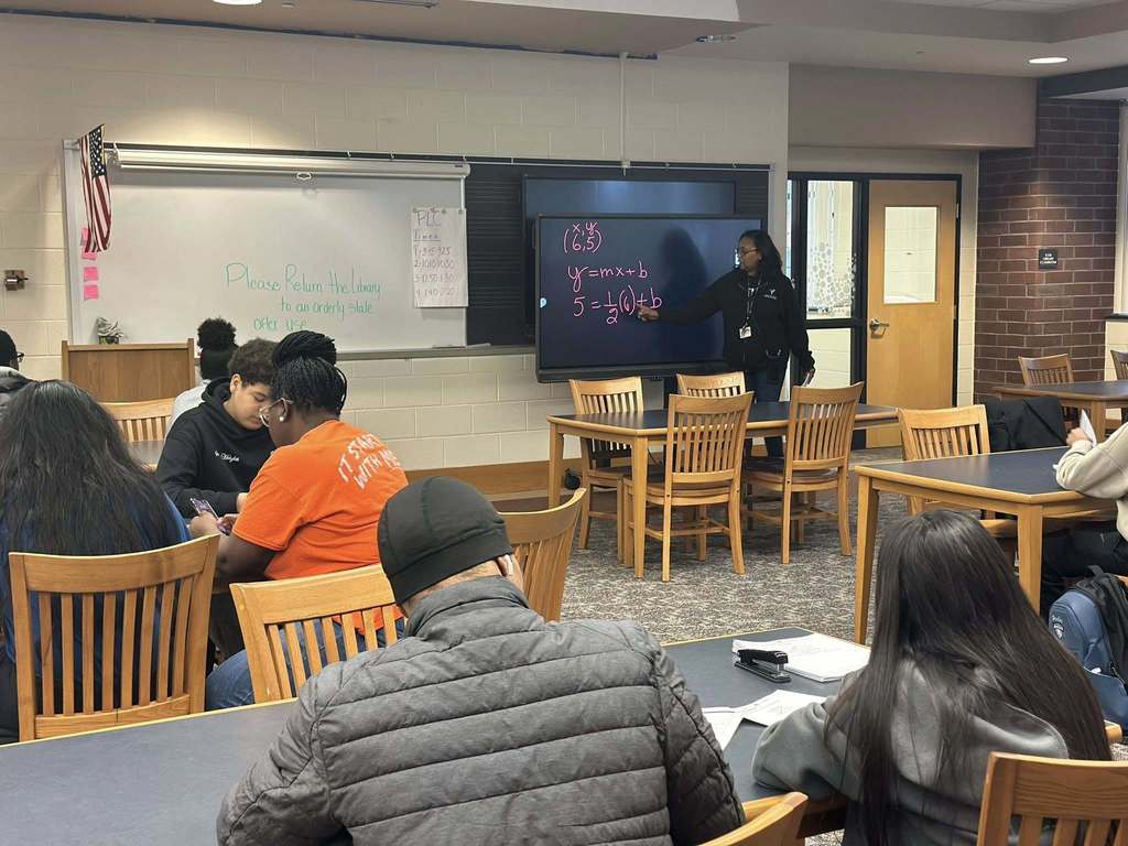 A district staff member is pointing to a smart board that has algebra on it in a school library. Students are sitting at tables observing and taking notes.