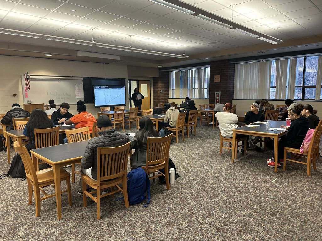 A school library filled with students who are sitting at tables, focusing on a district staff member who is standing near a smart board displaying a graph.