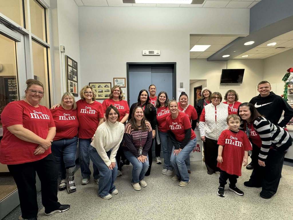 A group of district staff members and one child are posing indoors in a lobby area, most of them wearing red "Davis m&m" shirts.