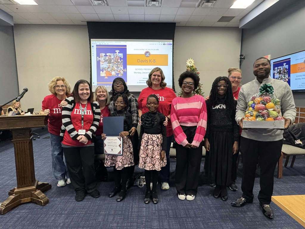 A group of district staff members and children are standing in a conference room. Some district staff members are wearing red "Davis m&m" shirts . A man is holding a colorful gift basket. A presentation screen in the background reads "Davis K-8."
