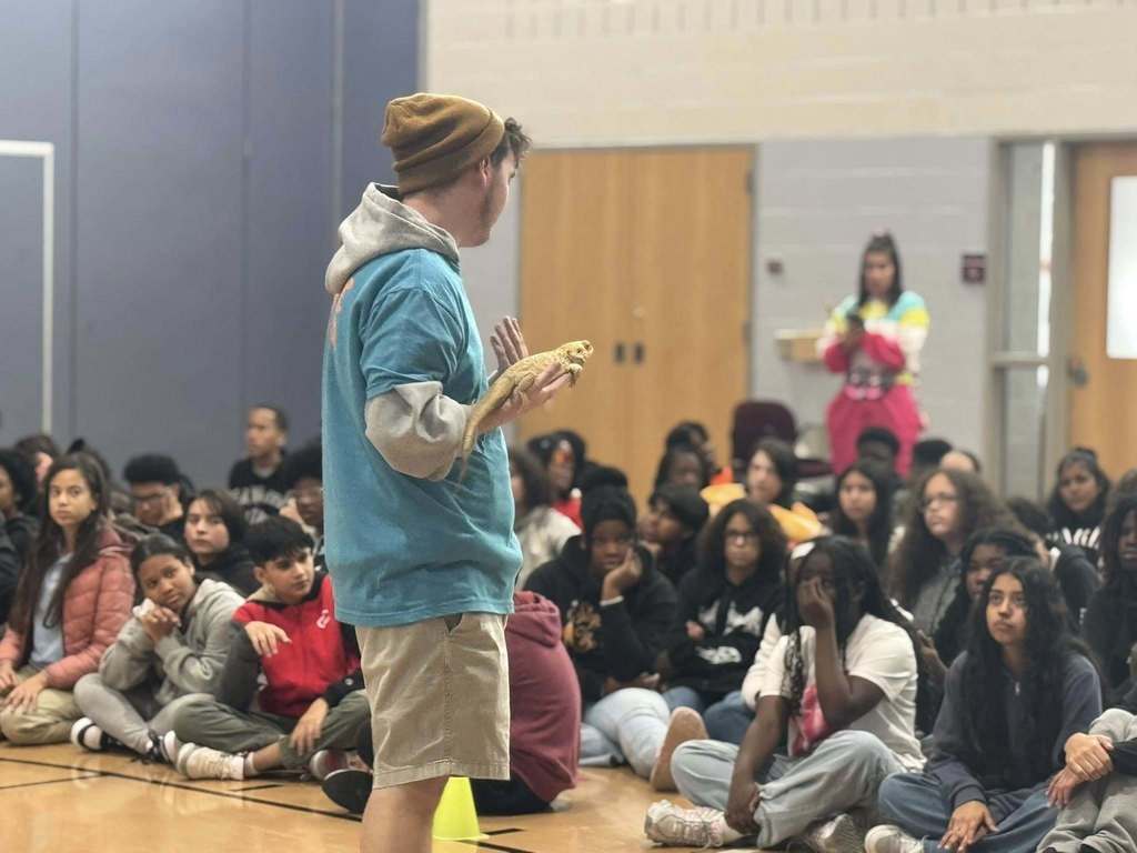 A person in a beanie and blue hoodie is holding an animal in front of a large group of students who are seated in a school gym.