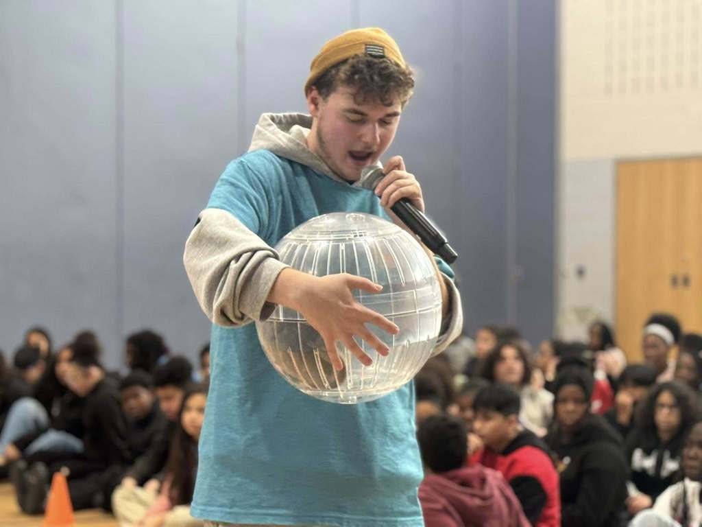 A person in a beanie and blue hoodie is holding a clear, inflatable globe with an animal inside of it while speaking into a microphone. He is standing in front of a large group of students who are seated in a school gym.