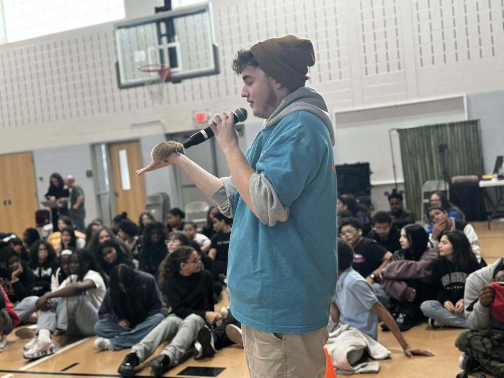 A person in a beanie and blue hoodie is holding a microphone in one of his hands and an animal in his other hand while a large group of students are seated in a school gym.