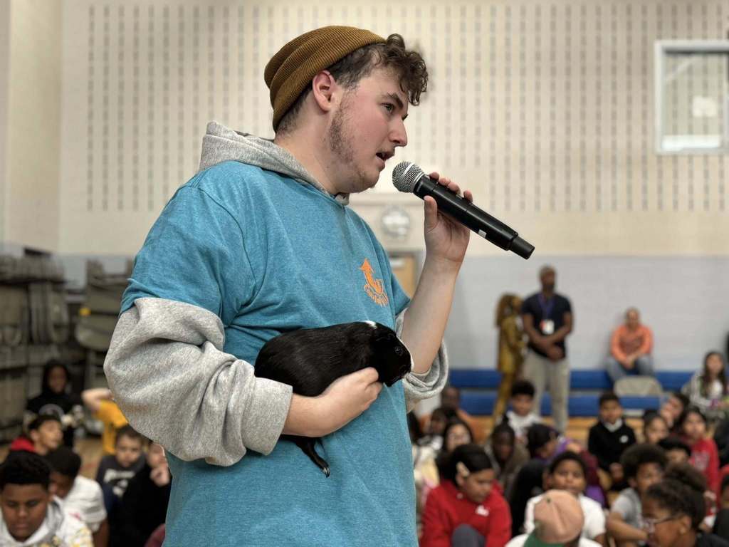 A person in a beanie and blue hoodie is holding a microphone in one of his hands and an animal in his other hand while a large group of students are seated in a school gym.