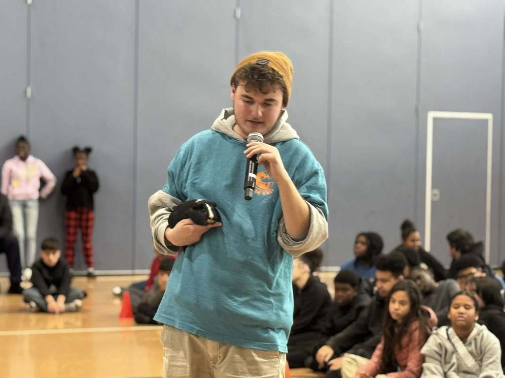 A person in a beanie and blue hoodie is holding a microphone in one of his hands and an animal in his other hand while a large group of students are seated in a school gym.