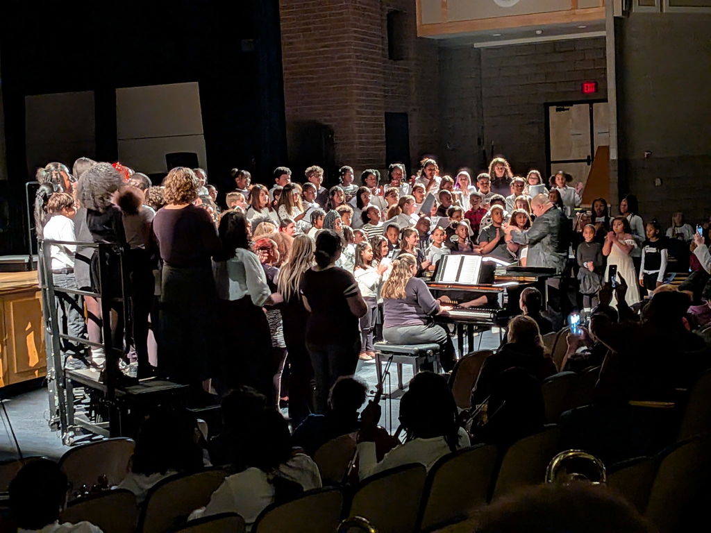 A large choir of students are singing on a dimly lit stage, facing a conductor in a concert setting. A pianist accompanies them as an audience of people are watching attentively.