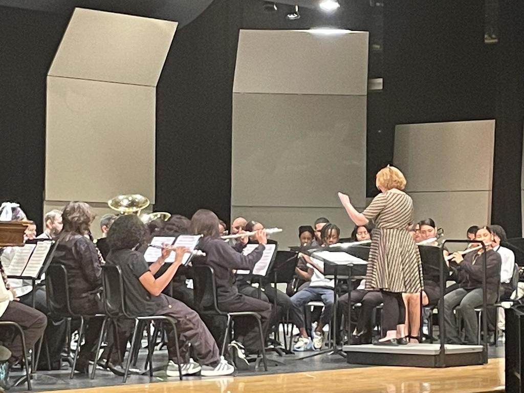 A conductor is leading a group of students who are playing wind instruments on stage in a concert setting.