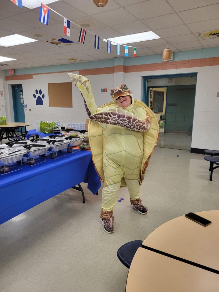 A person dressed in a turtle costume has their hands in the air while standing in a school cafeteria with a blue tablecloth-covered table holding food trays. Festive decorations are hanging from the ceiling.