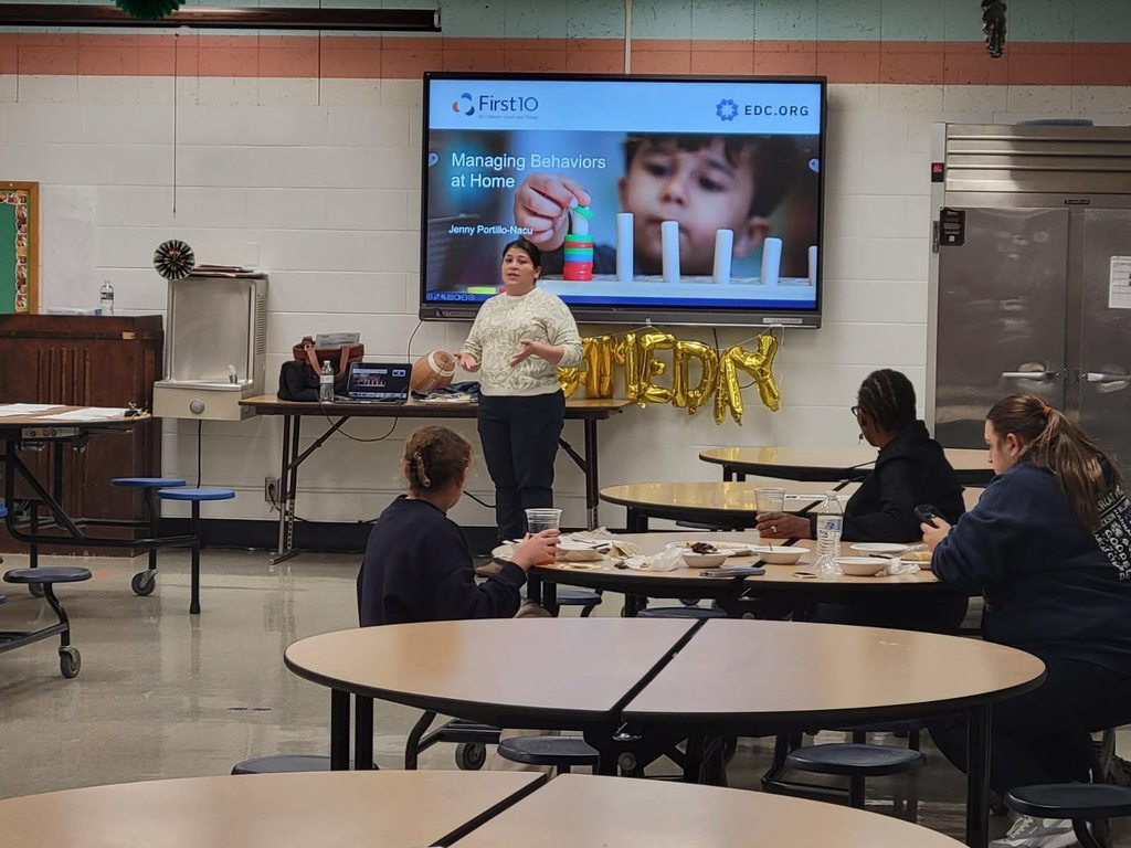 A woman is presenting on "Managing Behaviors at Home" in a school cafeteria, with a group of adults seated at round tables observing her.