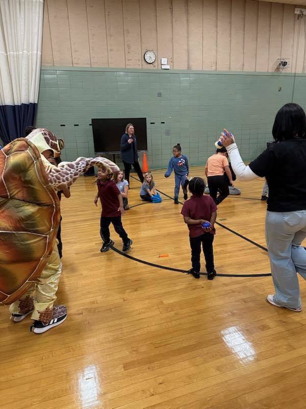 A person in a turtle costume is interacting with excited children who are playing in a school gym.