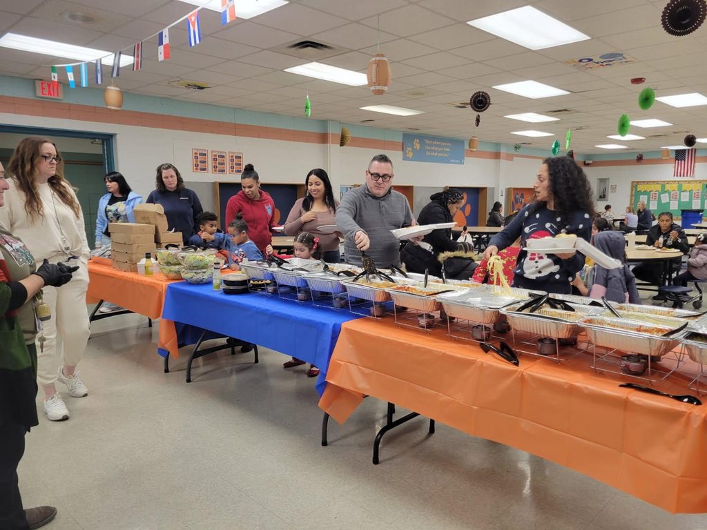 A group of people are using utensils to pick up food at a school event in a school cafeteria. Tables are covered in blue and orange tablecloths, with festive decorations hanging from the ceiling.