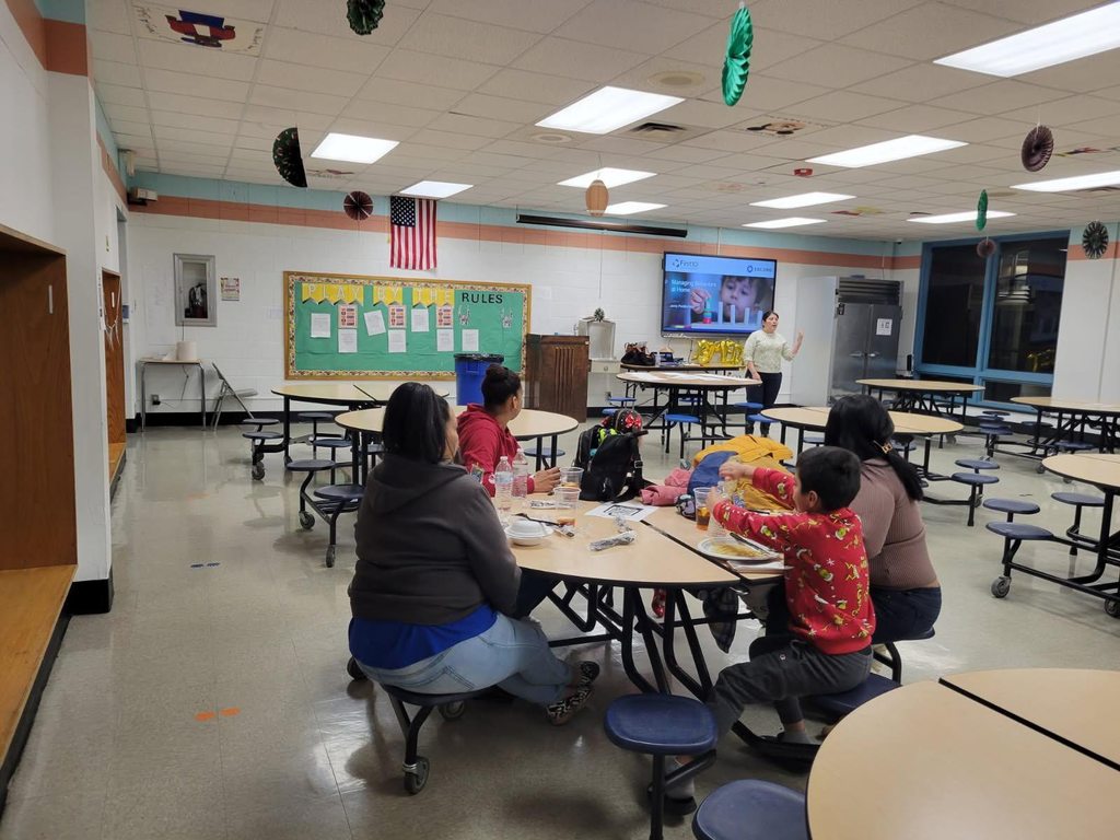 A woman is presenting on "Managing Behaviors at Home" in a school cafeteria, with a group of adults and kids seated at round tables observing her.