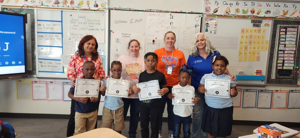 A group of five students are holding certificates in a school classroom while three district staff members are standing with them.