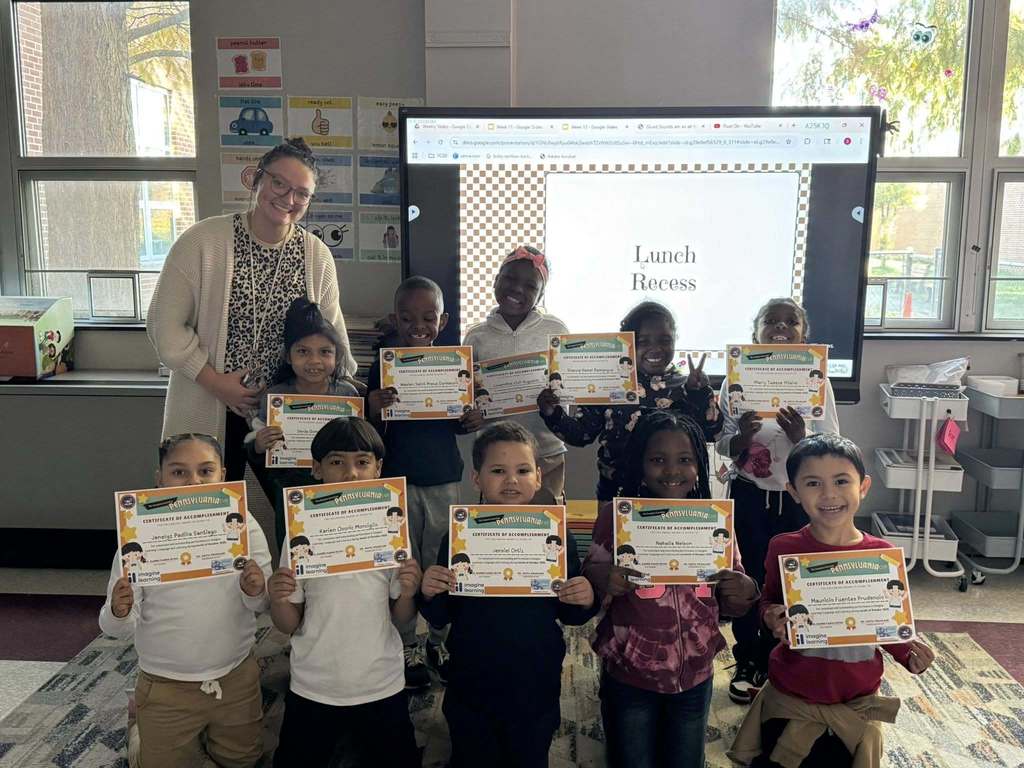 A district staff member is posing with a group of ten students who are holding certificates in a school classroom. A "Lunch Recess" slide is displayed behind them.