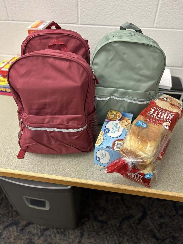 Two backpacks, one red and one mint green, sit on a table with a loaf of white bread and food items, suggesting a prepared meal or donation.