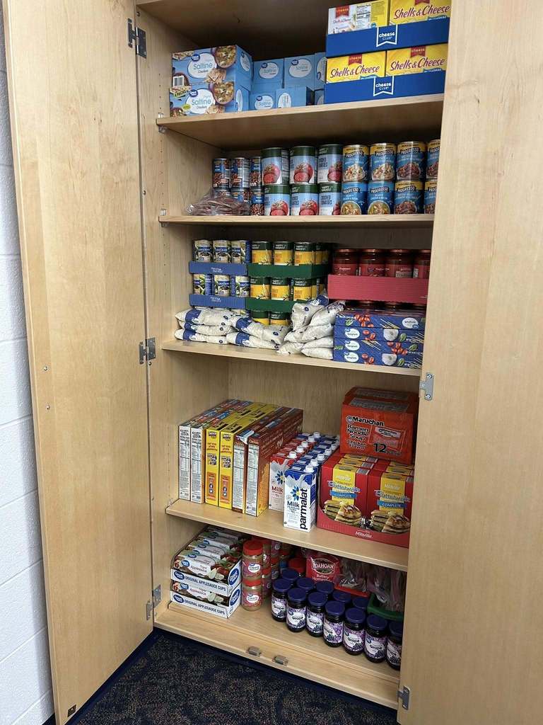 A wooden pantry filled with various boxed and canned foods, including mac and cheese, cereal, canned vegetables, rice, instant noodles, and jars of jam.