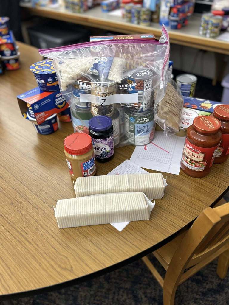 A table filled with various pantry items, including jars of peanut butter and jelly, cans, pasta sauce, and crackers, suggesting food donation prep.