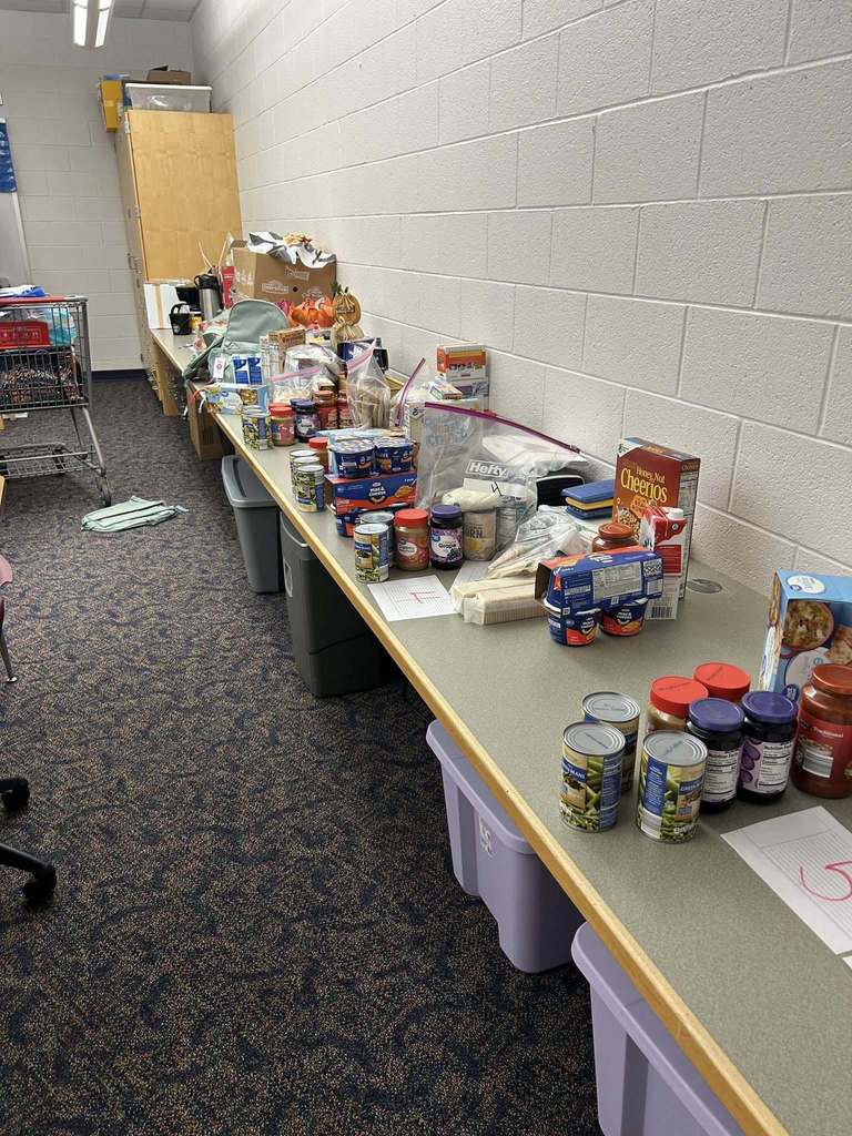 A long table against a wall holds various food items, including canned goods, cereals, and jarred sauces, arranged for donation. The room has a tidy, organized feel.