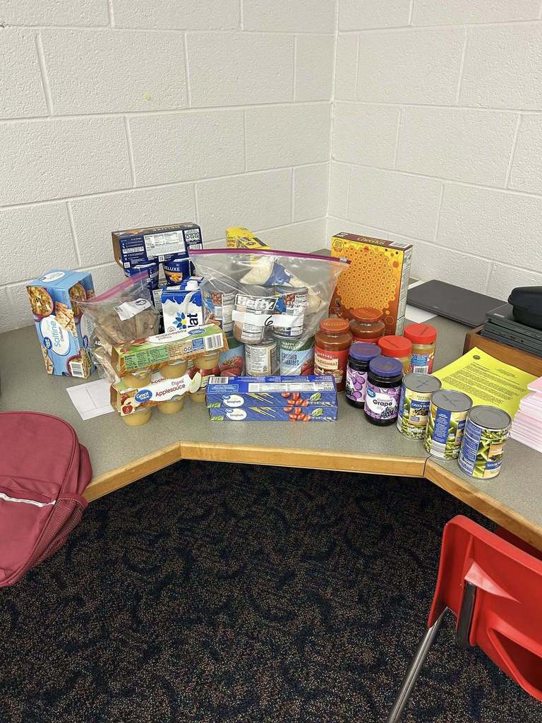 A collection of canned and packaged food items, including pasta, sauce, jam, and snacks, is arranged on a corner desk. A red backpack and a red chair are nearby.