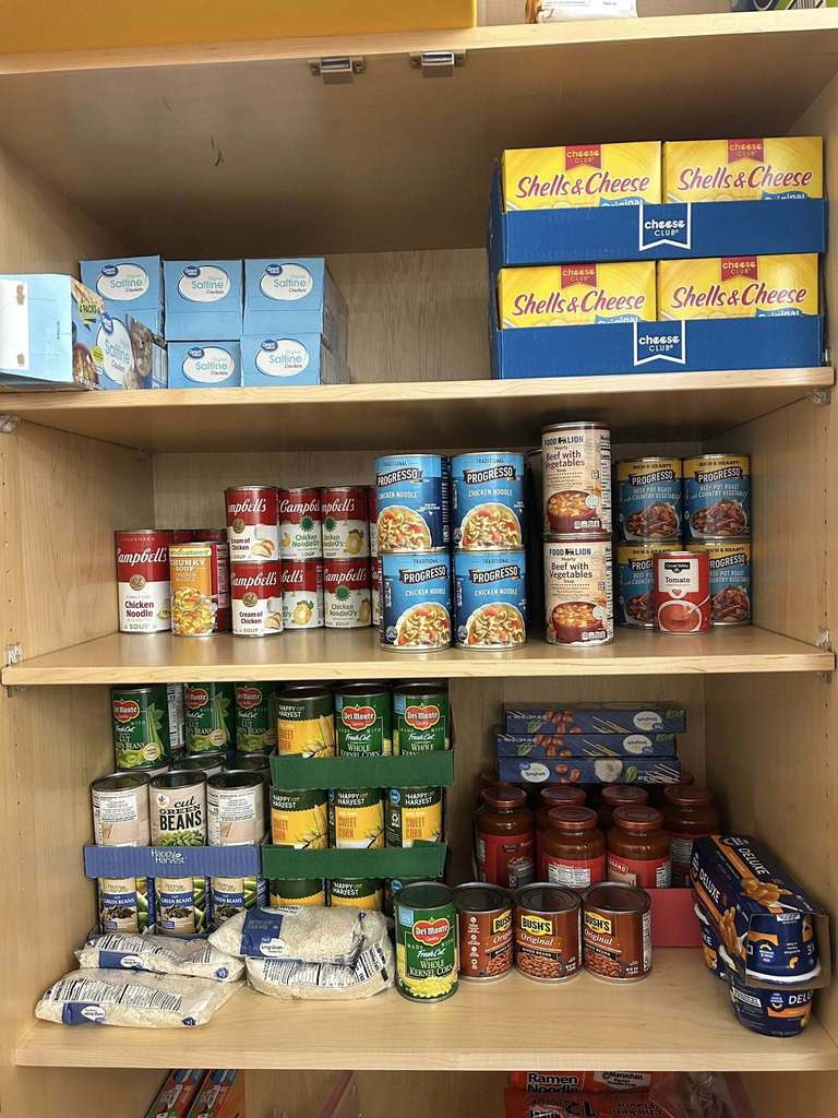 A neatly organized pantry with boxed mac and cheese, canned soups, vegetables, and sauces. Rice and beans are on lower shelves.