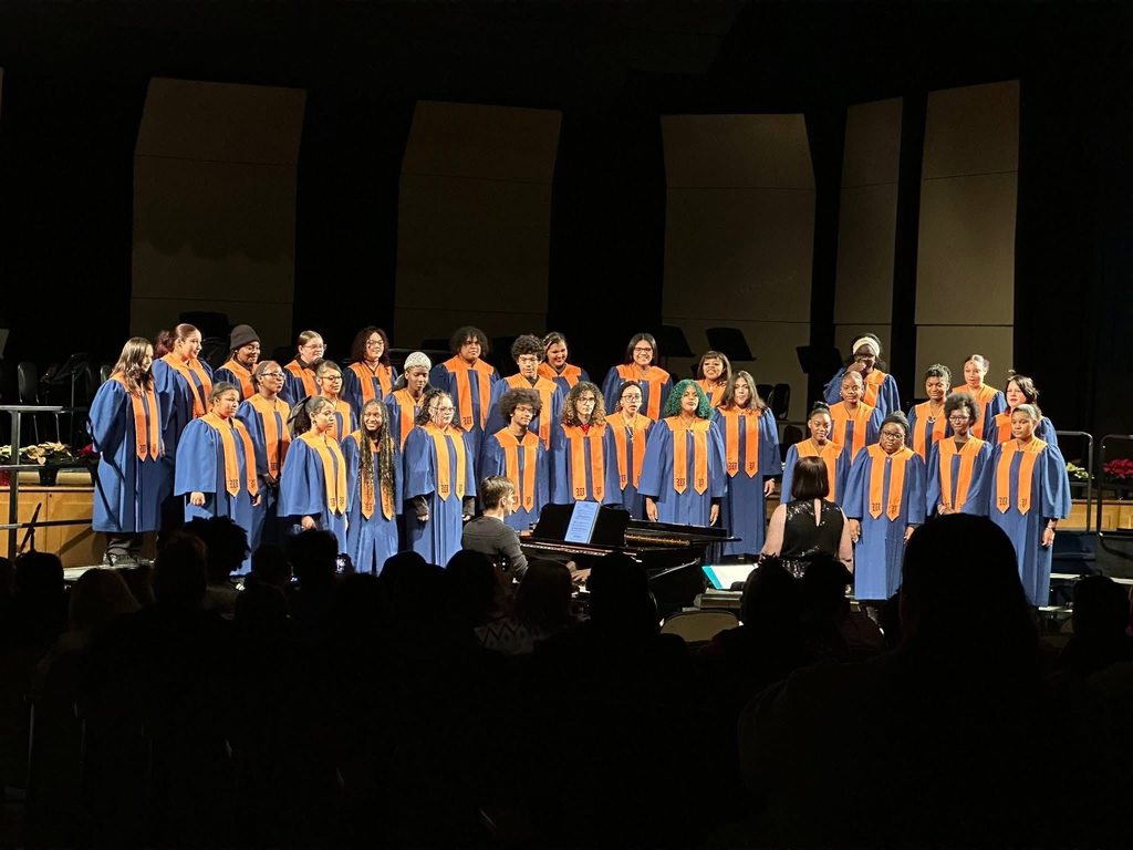A choir of 30 singers stands on stage in blue robes with orange stoles, singing in a concert setting. A conductor and pianist accompany them.