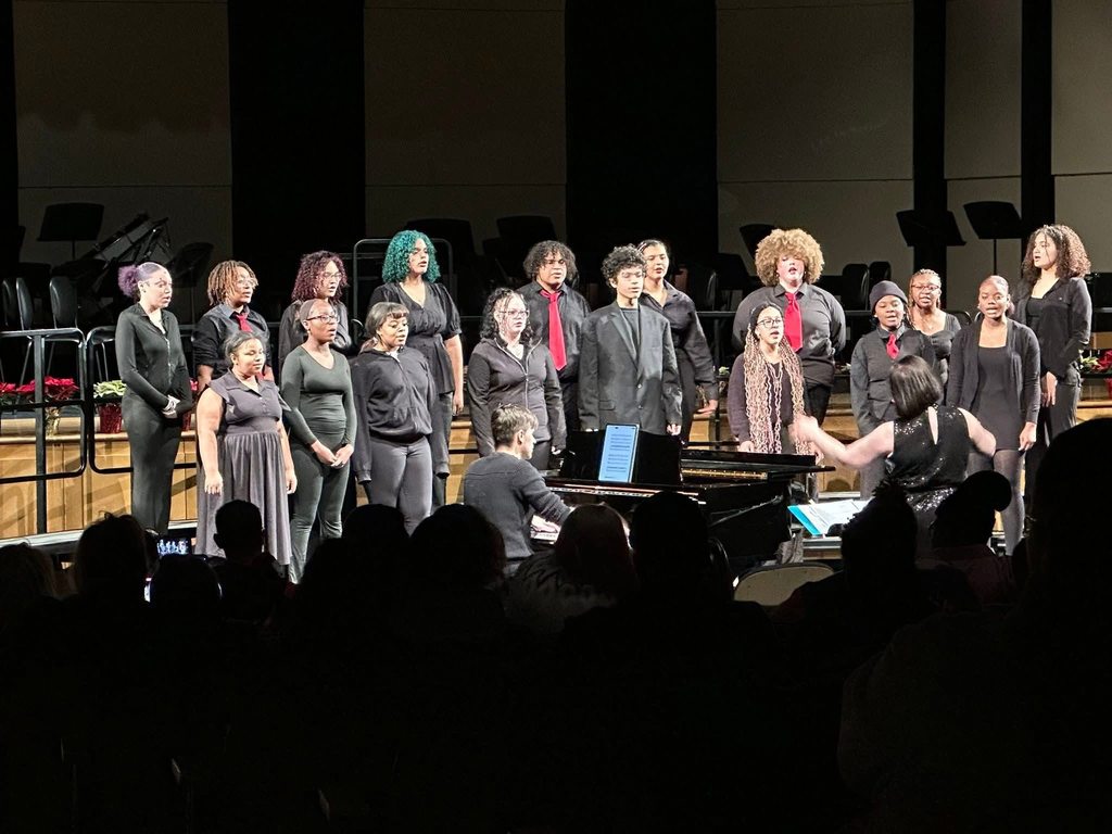 A choir composed of fourteen individuals dressed in black attire are performing on stage in a concert setting. They're standing in rows, singing with a conductor and pianist.