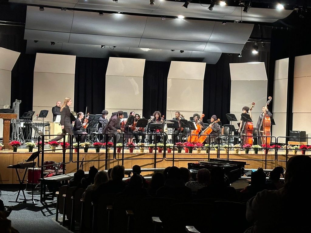 A symphony orchestra on stage, musicians with string instruments, a conductor in motion in a concert setting. Red poinsettias line the stage for a festive mood.
