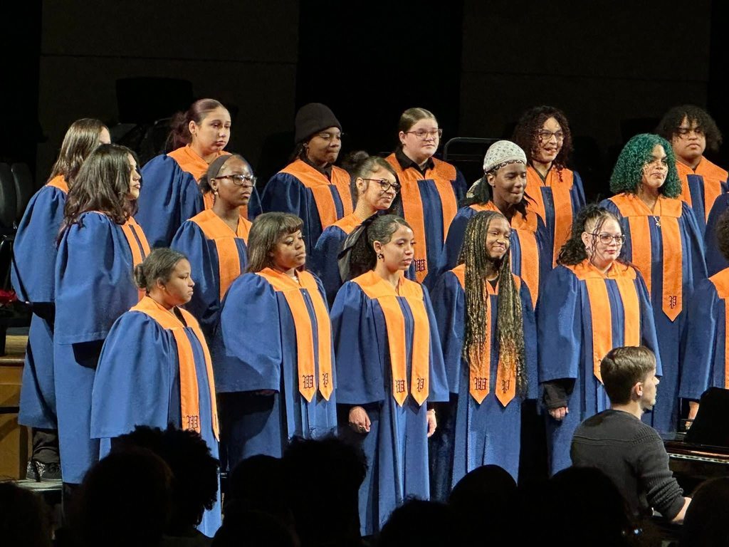 A choir of individuals in blue and orange robes are singing on stage in a concert setting. A pianist accompanies them.