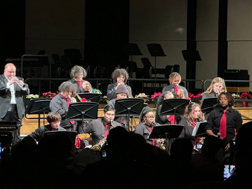 A jazz band performs on stage, featuring a group of musicians wearing black with red ties in a concert setting. They are playing various instruments.