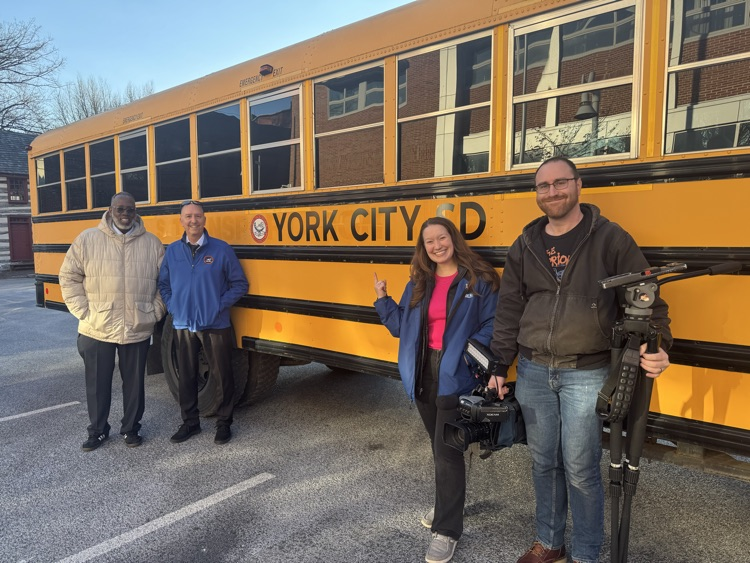 Four people, including district staff members and a news team are standing in front of a yellow school bus with "York City SD" on its side.