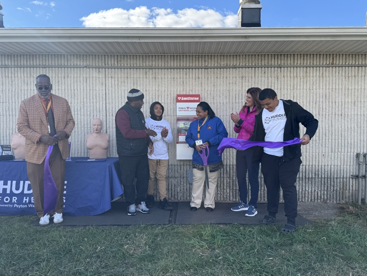 District staff members and community members are standing near a SaveStation machine and a mannequin, cutting a ribbon.