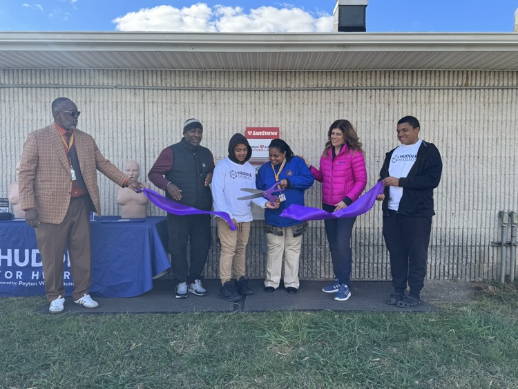 District staff members and community members are standing near a SaveStation machine and a mannequin, cutting a ribbon.