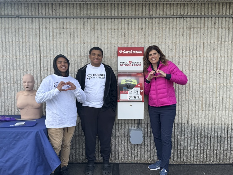 A woman and two boys are standing near a SaveStation machine and a mannequin, with one of the boys and the woman doing a heart gesture.