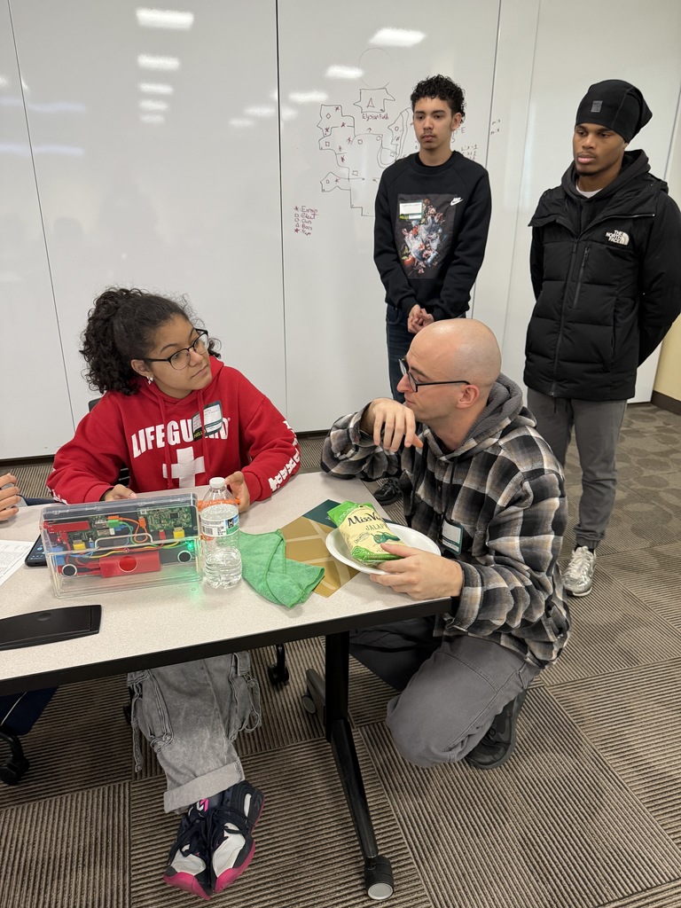 A student is sitting at a table with electronics and a water bottle, engaging in a discussion with a district staff member. Two students can be seen standing behind them in the background.