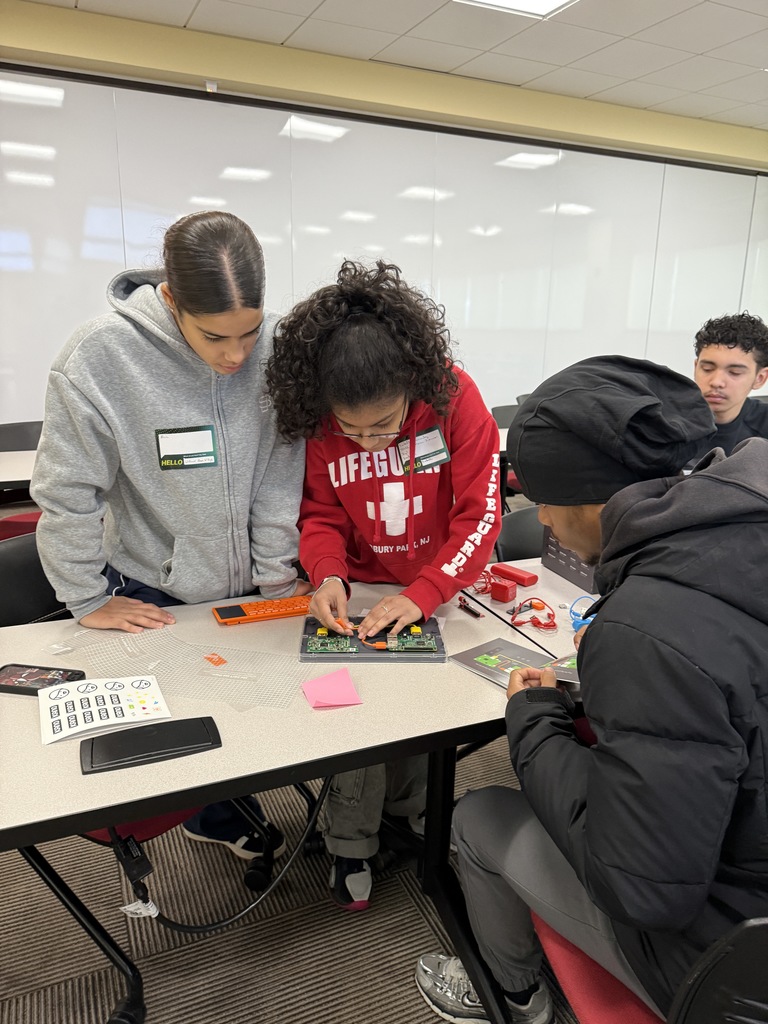 A group of three students are collaborating on a tech project, examining circuit boards on a table. Another student can be seen nearby.