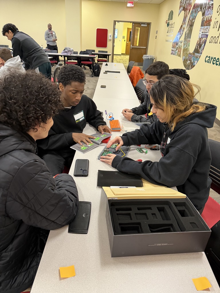 A group of four students are collaborating on a tech project, examining circuit boards on a classroom table. Other people can be seen in the background. 