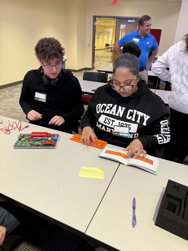 Two students are collaboratively assembling a circuit board at a table. One of the students is reading a manual while the other is observing.