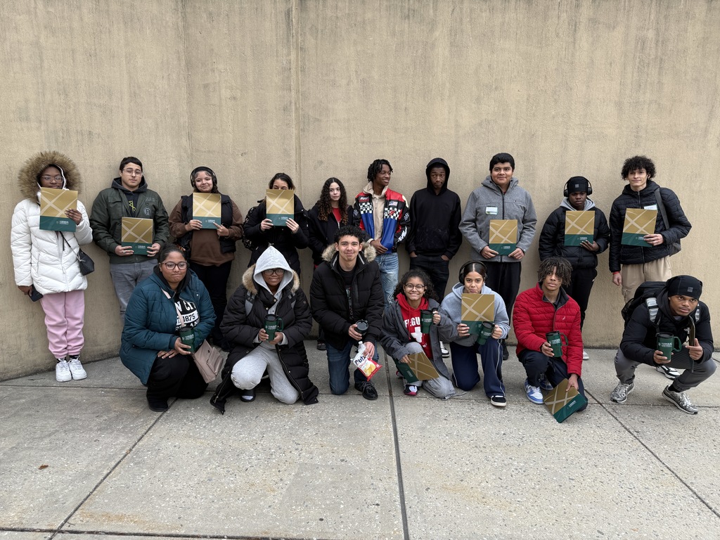 A group of students are holding gold and green boxes while standing outside in front of a concrete wall. 