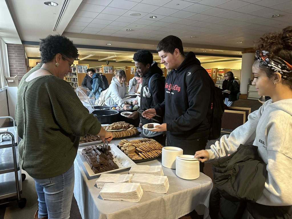 A district staff member is serving a group of students at a buffet table with cookies, brownies, and other snacks in a school library.