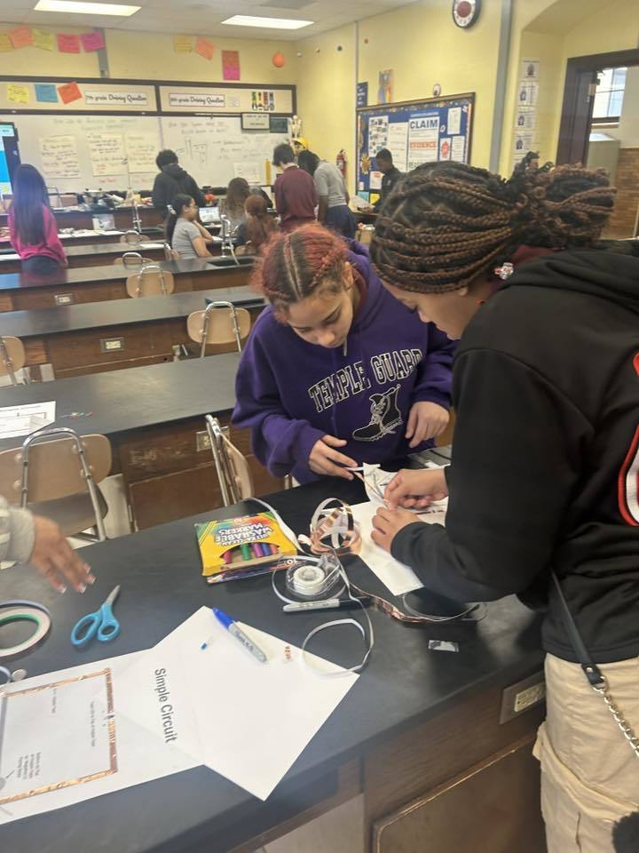Two students are working together on a simple circuit project in a school classroom. Tools, papers, and craft supplies cover the table. Other people can be seen in the background.
