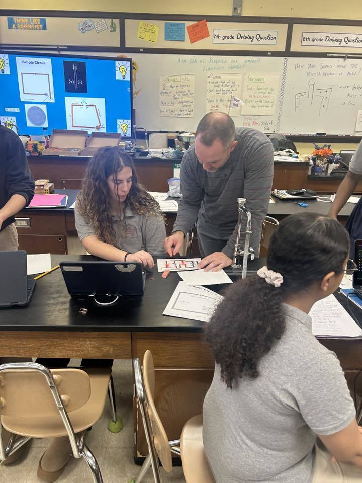 A district staff member is leaning over to help a student with a circuit diagram in a school classroom. Other people can be seen nearby.