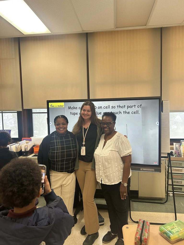 Dr. Andrea Berry-Brown, Superintendent of Schools, a district staff member and a community member are standing in front of a digital whiteboard. A student can be seen in the foreground taking a photo of them with a phone.