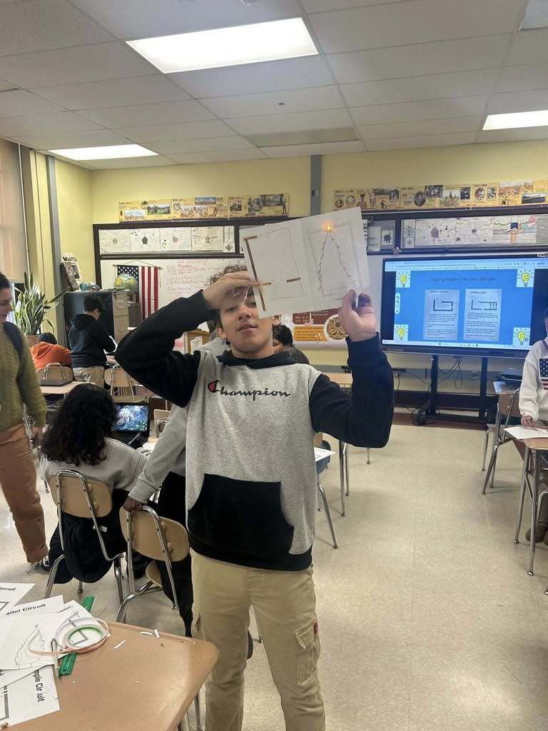 A student is standing in a school classroom, holding up a paper circuit with a glowing LED. Other people can be seen in the background.