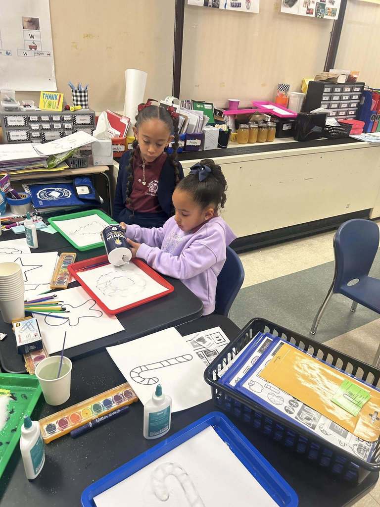 Two young students are engaging in an art project in a school classroom. One of the students is pouring white salt onto a cookie cutter shape, while the other student is observing her. Art supplies and papers are covering the table.