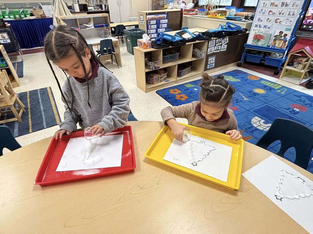 Two young students are using plastic cups to trace star shapes on paper in a school classroom.